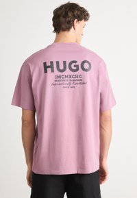 Ružový bavlnený tričko s logom "HUGO" v tmavošedej farbe, s textom MCMXCIII a "zaregistrovaná ochranná známka" na chrbte. Krátke rukávy a voľný strih.