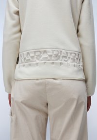 Pullover in pile beige con una fascia in rilievo e testurizzata all'orlo. Il tessuto appare morbido con una lucentezza leggera e sottile. Pantaloni color beige chiaro.