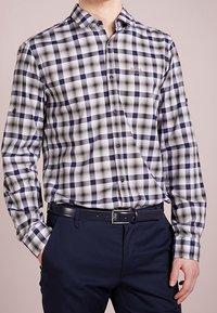 Mężczyzna w czarno-biało-beżową koszulę w kratę zapinaną na guziki, włożoną w granatowe spodnie z czarnym paskiem.