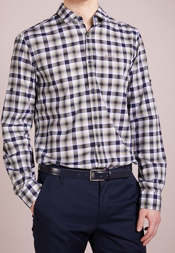 Mężczyzna w czarno-biało-beżową koszulę w kratę zapinaną na guziki, włożoną w granatowe spodnie z czarnym paskiem.