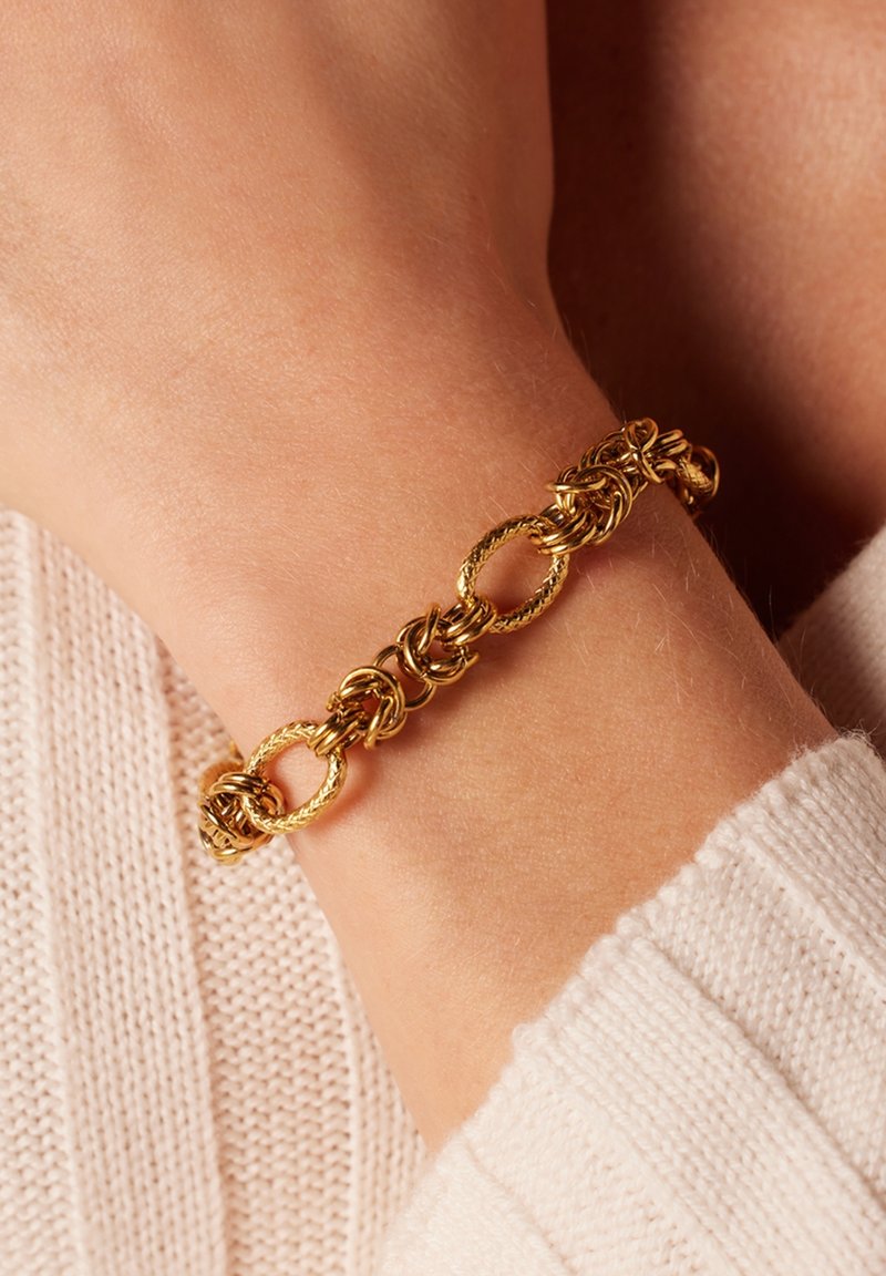 Gouden armband met een ontwerp van onderling verbonden kettingen en getextureerde elementen. Draagt op een pols, tegen een achtergrond van licht gebreide stof.
