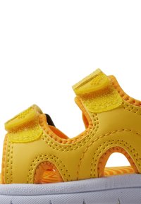 Gule sandaler med en teksturert gummioverdel, som har to justerbare borrelåsfester, og en hvit såle med en mønstrete fotbed.