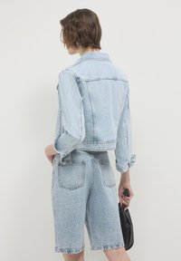 Persona con capelli corti castani che indossa una giacca di denim azzurro chiaro e pantaloni corti di denim a vita alta abbinati, mentre tiene una piccola borsa nera.