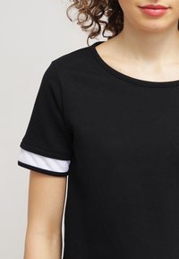 Osoba s kudrnatými vlasy, oblečená v černém tričku s krátkým rukávem a bílým detailem na manžetách, na pozadí s jednobarevným světlým pozadím.