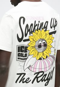 Valkoinen puuvillainen t-paita, jossa on painettu kuva aurinkolaseja käyttävästä hymyilevästä auringonkukasta ja teksti "Sunnie’s Soaking Up The Rays".