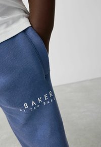 Sinised dressipüksid, mis on valmistatud pehmest kangast. Püksil on reie peal suur valge logo, millel on kiri "BAKER BY TED BAKER". Nähtav õrn tekstuur.