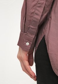 Chemise à manches longues avec boutons, en tissu mauve présentant une texture lisse, bouton blanc au poignet et fentes latérales près de l'ourlet.