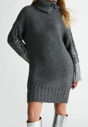 Femme portant une robe pull tricotée gris foncé avec des manches torsadées et un col montant avec un bouton, associée à des bottes argentées montantes.