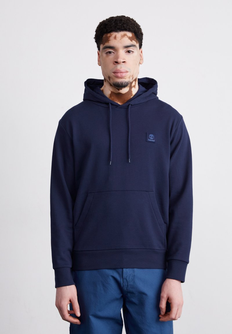 Dunkelblaue Baumwoll-Hoodie mit einer Vordertasche, Kapuze mit Kordelzug und einem kleinen Logopatch. Verfügt über gerippte Bündchen und Saum. Kombiniert mit blauen Hosen.