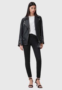 Veste de motard en cuir noir avec des fermetures éclair argentées, portée sur un t-shirt blanc et associée à un jean skinny noir et des sandales à talons.