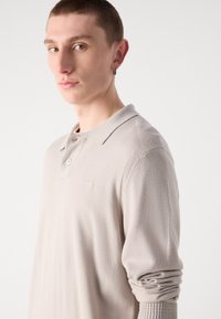 Beige långärmad polo skjorta med krage, med en diskret logga på bröstet, texturerade ärmar och en figurnära design.