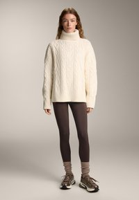 Crème oversized kabelgebreide trui met een hoge kraag, gecombineerd met donkerbruine legging en sportieve schoenen met grijze accenten.