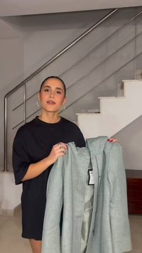 Vrouw met donker haar in een zwarte jurk houdt een grijze jas met labels vast in een modern huis bij een trap met metalen leuning.