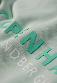 Vaaleanvihreä kangas, jossa koholla olevaa tekstiä tummanvihreänä ja valkoisena. Pinta on hieman sileä ja siinä on kudottu kuvio.