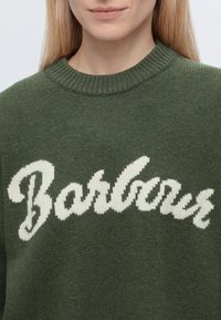 Femme portant un pull en tricot vert avec le mot "Barbour" en grandes lettres blanches cursives sur le devant.