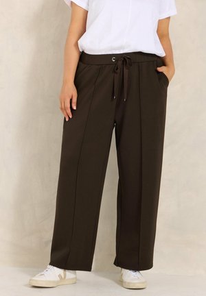 Persoon draagt losse donkerbruine wijde broek met trekkoord in de taille, wit shirt met korte mouwen en witte sneakers, staand tegen een effen achtergrond.