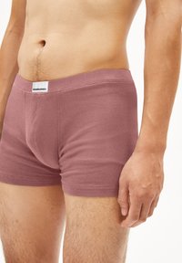 Homme portant un boxer mauve, se tenant debout avec le bras droit détendu le long du corps, torse et jambes visibles sur un fond blanc.