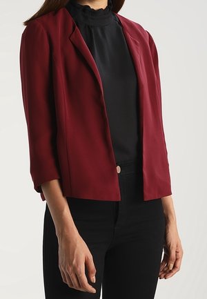 Femme portant un blazer bordeaux court sur un chemisier noir à col montant et un pantalon noir, debout avec les bras détendus le long du corps.