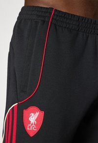 Schwarze Jogginghose mit strukturiertem Oberflächen, versehen mit einem roten L.F.C.-Logo und vertikalen roten Streifen an den Seiten. Elastischer Bund und Taschen.