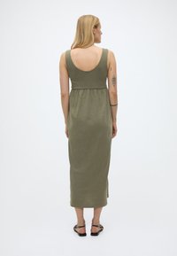 Γυναίκα με ξανθά μαλλιά, φοράει αμάνικο μεσομήκες φόρεμα ελιάς και μαύρα σανδάλια, εμφανιζόμενη από την πλάτη σε ουδέτερο φόντο.