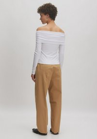Weißes Off-Shoulder-Oberteil mit langen Ärmeln und gerafftem Design. Kombiniert mit lockeren, hellbraunen Hosen und schwarzen Fransen-Schuhen.