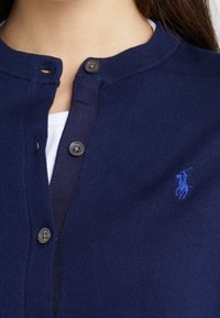 Mörkblå cardigan med rund halsringning, knappstängning och en liten broderad logotyp på bröstet. Tillverkad av ribbad stickad tyg.