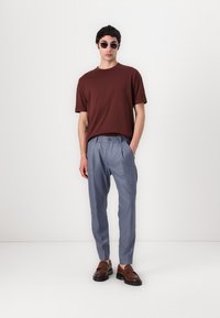 Bruine korte mouwen t-shirt, lichtblauwe broek met plooien, en bruine instappers. Accessoires zijn ronde zonnebrillen. Minimalistisch ontwerp.