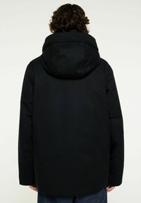 Person med lockigt hår som bär en svart huvtröja, sedd från ryggen mot en enkel ljus bakgrund.