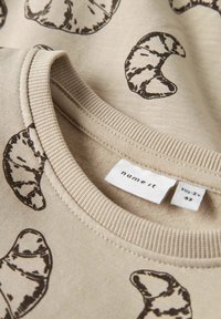 Sweatshirt beige avec encolure ronde, ornée de motifs de croissants marron. Étiquette visible affichant la marque "name it" et la taille 1½-2Y. Texture douce.