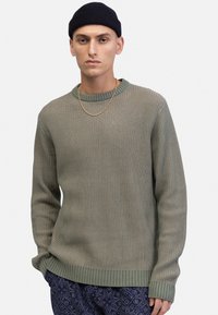 Grüner gerippter Strickpullover mit rundem Ausschnitt und langen Ärmeln. Leichtes Material mit strukturierter Oberfläche. Kombiniert mit gemusterten blauen Hosen.