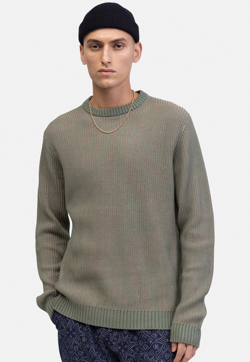 Grüner gerippter Strickpullover mit rundem Ausschnitt und langen Ärmeln. Leichtes Material mit strukturierter Oberfläche. Kombiniert mit gemusterten blauen Hosen.