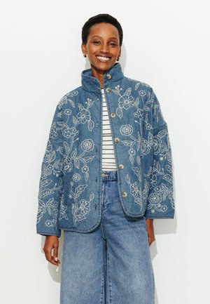 Femme souriante portant une veste matelassée bleue avec broderie florale blanche, chemise rayée et jean large en denim sur un fond uni.