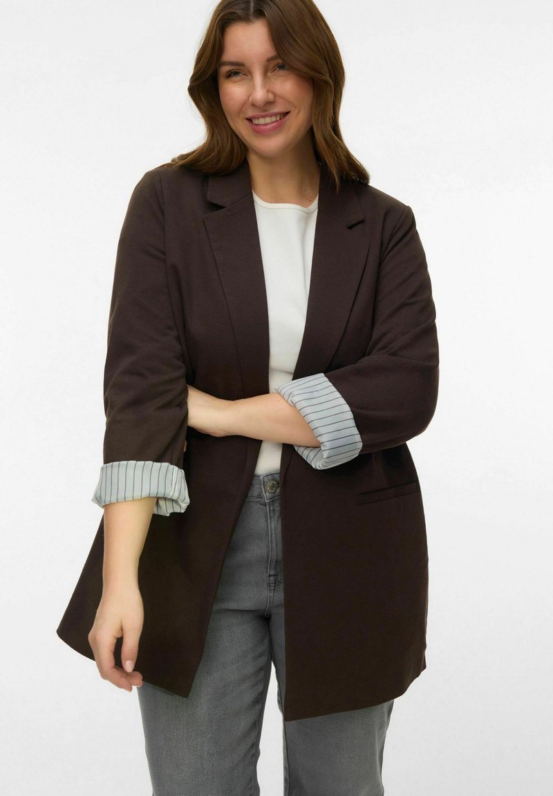 Blazer marron avec col cranté et poignets rayés retroussés. Porté par-dessus un haut blanc, associé à un jean gris. Tissu lisse et coupe ajustée.