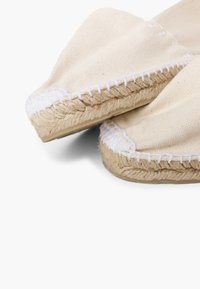 Canvas espadrilles met beige bovenkant, witte stiksels en een natuurlijke jute zool. Rond toelopend ontwerp met textielstof en gevlochten accenten.