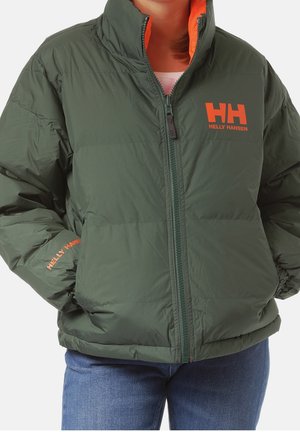 Personne portant une doudoune vert foncé avec des logos Helly Hansen orange sur la poitrine et la manche, associée à un jean bleu.