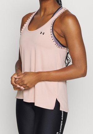 Persoon draagt een lichtroze mouwloos sporttopje van Under Armour over een donkere sportbeha en zwarte legging met Under Armour-logo.