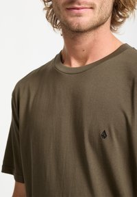 Olive green cotton t-shirt with a crew neck, short sleeves, and a small black logo on the chest. Smooth texture, relaxed fit.