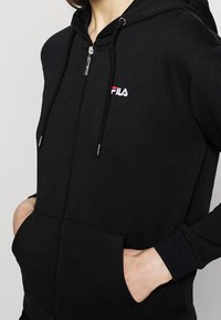 Sudadera negra con cremallera hecha de tela suave, que presenta un bolsillo frontal, cordones y un pequeño logotipo bordado en rojo y blanco en el pecho.