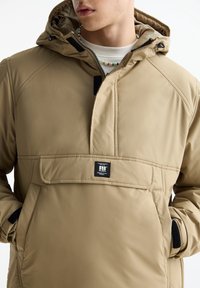 Beige geïsoleerd pulloverjack met hoge kraag. Voorzien van een zak aan de voorkant, ritssluiting en logopatch. Heeft een gladde textuur en een puffy ontwerp.