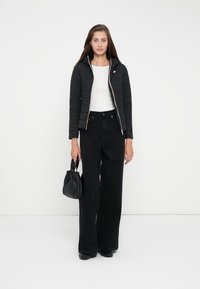 Quiltet svart jakke med glidelås og hette, kombinert med høyt liv og flagrende svarte jeans. Tilbehøret inkluderer en liten svart håndveske.