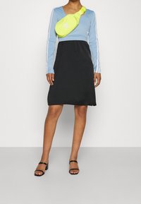 Camiseta de manga larga azul claro con rayas blancas, combinada con un vestido negro sin mangas. Riñonera amarilla con logotipo blanco, llevada con sandalias negras.