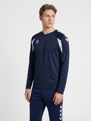 Jonge man die een marineblauw sportshirt met lange mouwen en bijpassende broek draagt met witte chevrondetails, kijkt opzij.