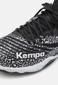 Fekete és fehér sportcipő, texturált hálós felsőrésszel, pöttyözött mintázattal, jellegzetes "Kempa" logóval és fekete cipőfűzőkkel.