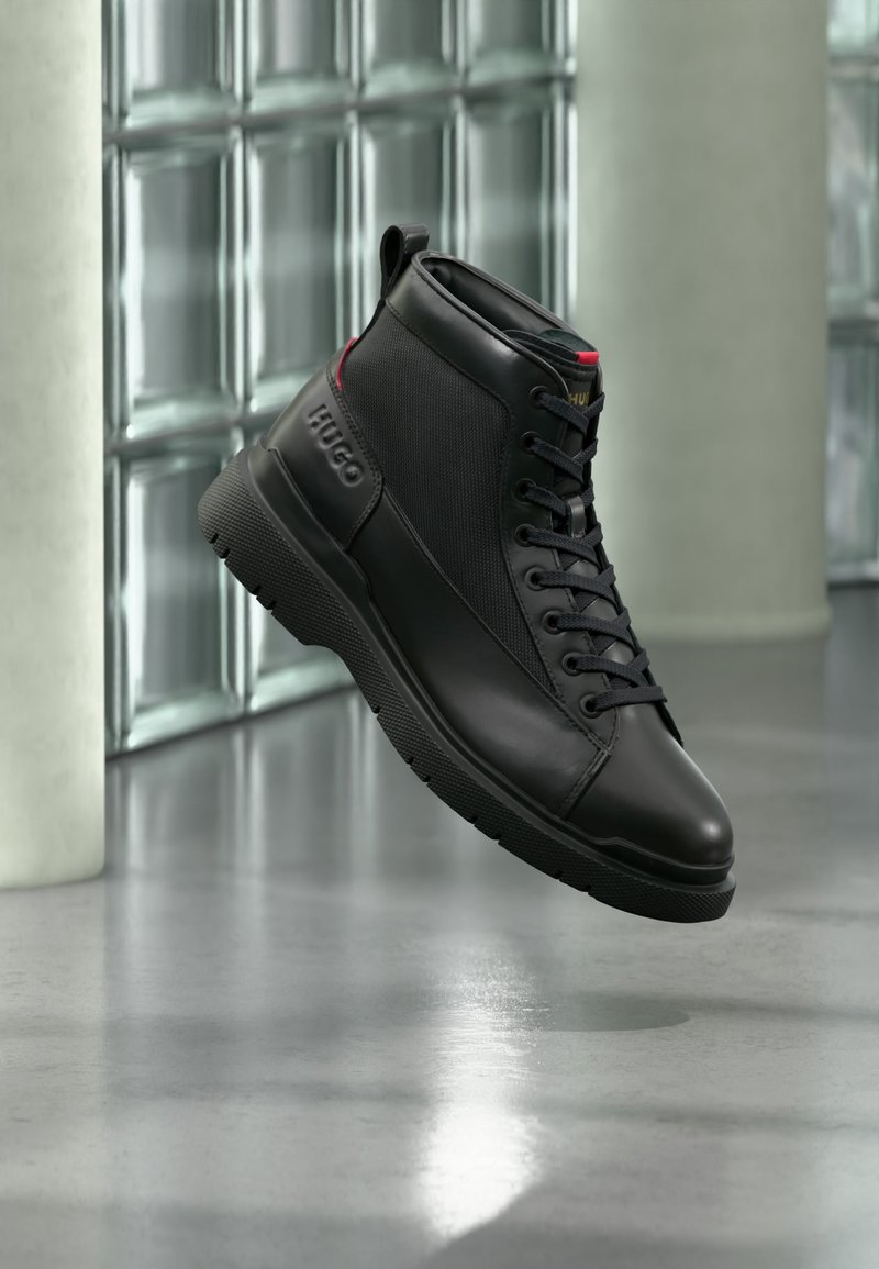 Sort højtop-sneakers med tekstureret sål og „HUGO“ branding på siden, vist mod en moderne glasblokvæg.