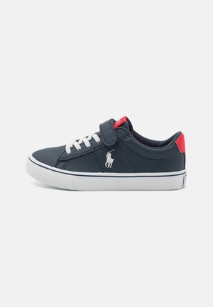 Baskets basses bleu marine avec lacets blancs, languette rouge au talon, bride velcro, semelle blanche, et un logo de joueur de polo blanc sur le côté.