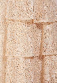 Beige spets tyg med blommönster, med lager av volanger som skapar en texturerad visuell effekt. Fina och detaljerade designelement är synliga.