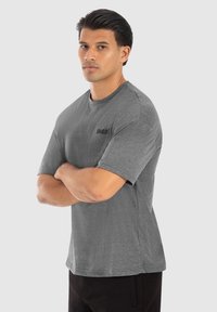 Camiseta gris de manga corta con cuello redondo, que presenta un pequeño logo negro en el lado izquierdo. La tela tiene una textura suave.