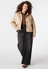 Lachende vrouw met krullend haar, gekleed in een beige jas, een bruine blouse met patroon, zwarte wijde jeans en bruine schoenen met stippen, staand tegen een witte achtergrond.