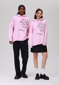 Svijetloružičaste oversized dukserice sadrže grafički tekst u crnoj boji. Modeli nose dukserice s crnim hlačama i suknjama, uparene s glomaznim crnim cipelama.