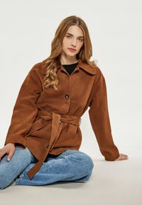 Giacca in finta pelle scamosciata marrone con colletto, chiusura frontale con bottoni e vita cintata, abbinata a jeans in denim azzurro chiaro.